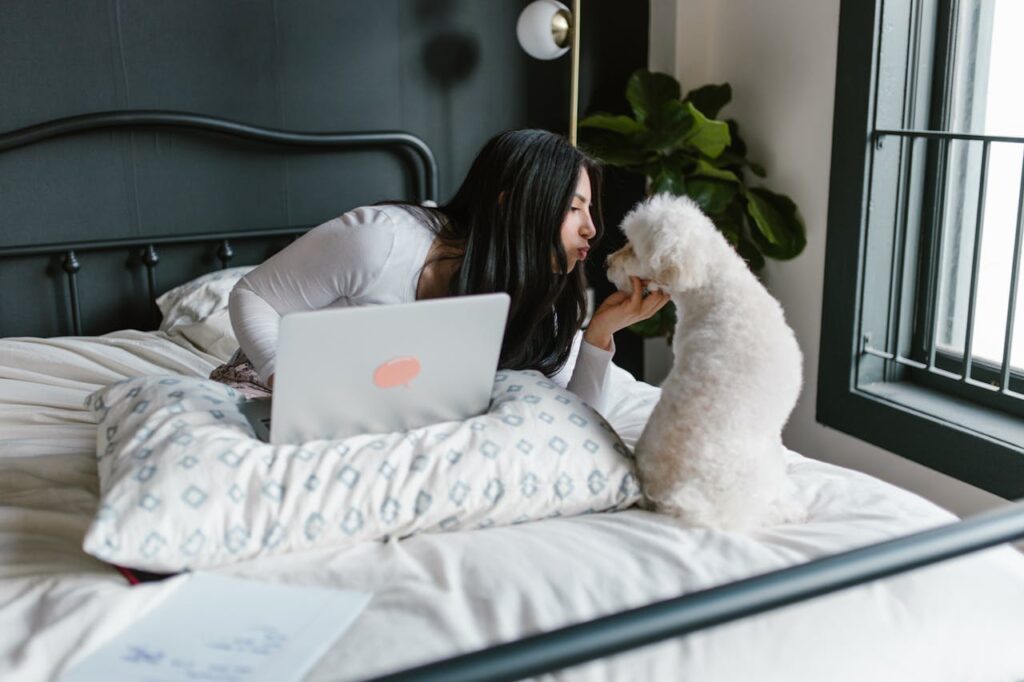 A woman kisses her dog on a bed while working remotely with a laptop, a blend of work and relaxation.